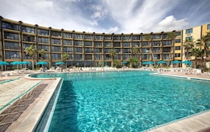A hotel with a large pool surrounded by lounge chairs and umbrellas, multiple floors with balconies, and a clear sky. - NEW Amazing Ocean Front Retreat in Daytona Beach! (Daytona Beach Shores)