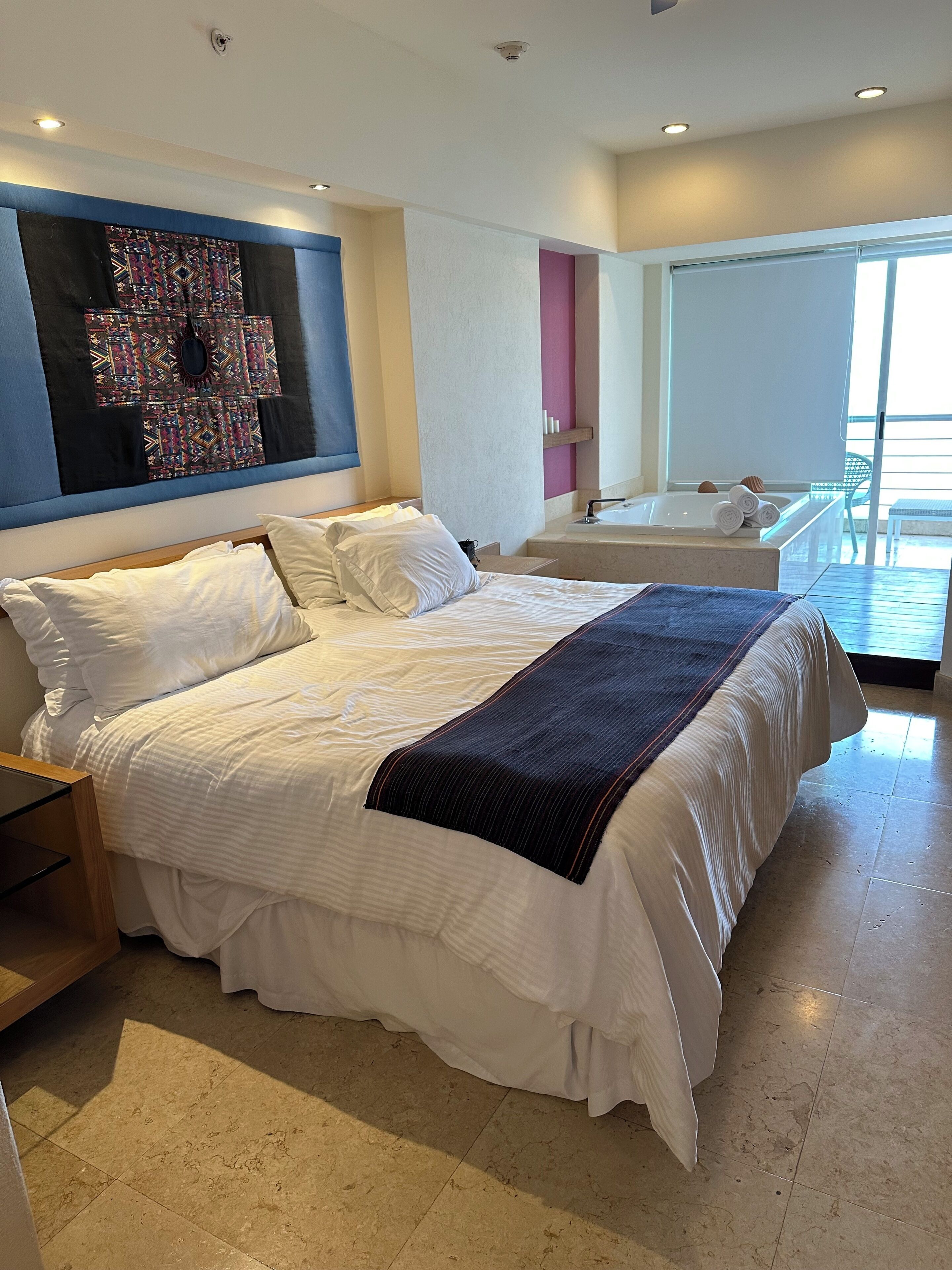 A modern hotel room with a large bed, a small sitting area with a bathtub, and a large window offering a view.