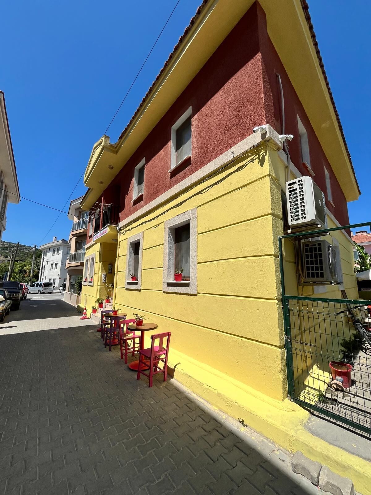 Un edificio amarillo con detalles rojos, un balcón y mesas exteriores con sillas rojas.