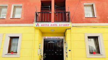 Un edificio de dos pisos con fachada amarilla, un balcón con barandilla negra y una puerta negra con un letrero encima.