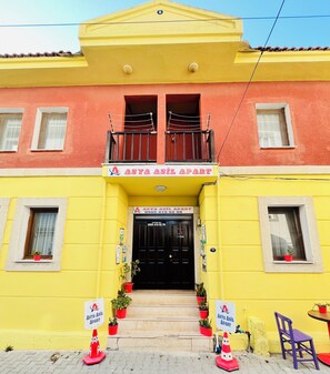 A two-story building with a yellow facade, a balcony with a black railing, and a black door with a sign above it.
