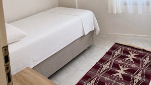 A small, single-bed room with a plain white bedspread, a pink pendant light, a sheer white curtain, and a patterned rug on the floor.