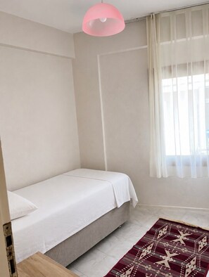 A small, single-bed room with a plain white bedspread, a pink pendant light, a sheer white curtain, and a patterned rug on the floor.