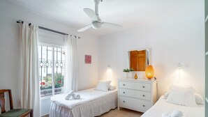 A hotel room with two beds, a ceiling fan, a window with curtains, a wooden door, a dresser with a mirror, and a small table with a lamp. - Holiday Home in Nerja With Sea Views & Pool (Nerja)