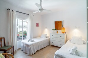 Ein Hotelzimmer mit zwei Betten, einem Deckenventilator, einem Fenster mit Vorhängen, einer Holztür, einem Schminktisch mit Spiegel und einem kleinen Tisch mit Lampe.