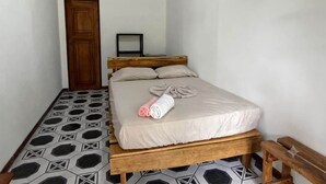 A single bed with a wooden frame, a headboard, and a white mattress. There is a wooden door to the left and a small framed picture on the wall.