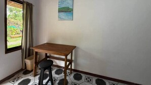 A small room with a wooden table and two stools, a window with curtains, and a painting on the wall.