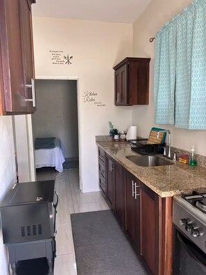A compact kitchen with granite countertops, a sink, and wooden cabinets. There are kitchen rules on the wall, a small window with patterned curtains, and a bed in the adjacent room.