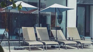 Zona de piscina con sillones de playa y sombrillas, con vista clara a la piscina y fachada de edificio moderna.
