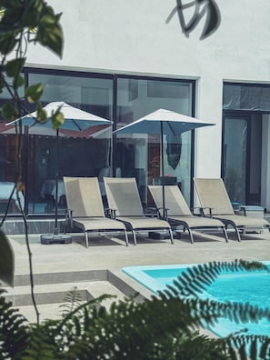 Ett område vid poolen med solstolar och parasoller, fri sikt över poolen och en modern byggnadsfasad.