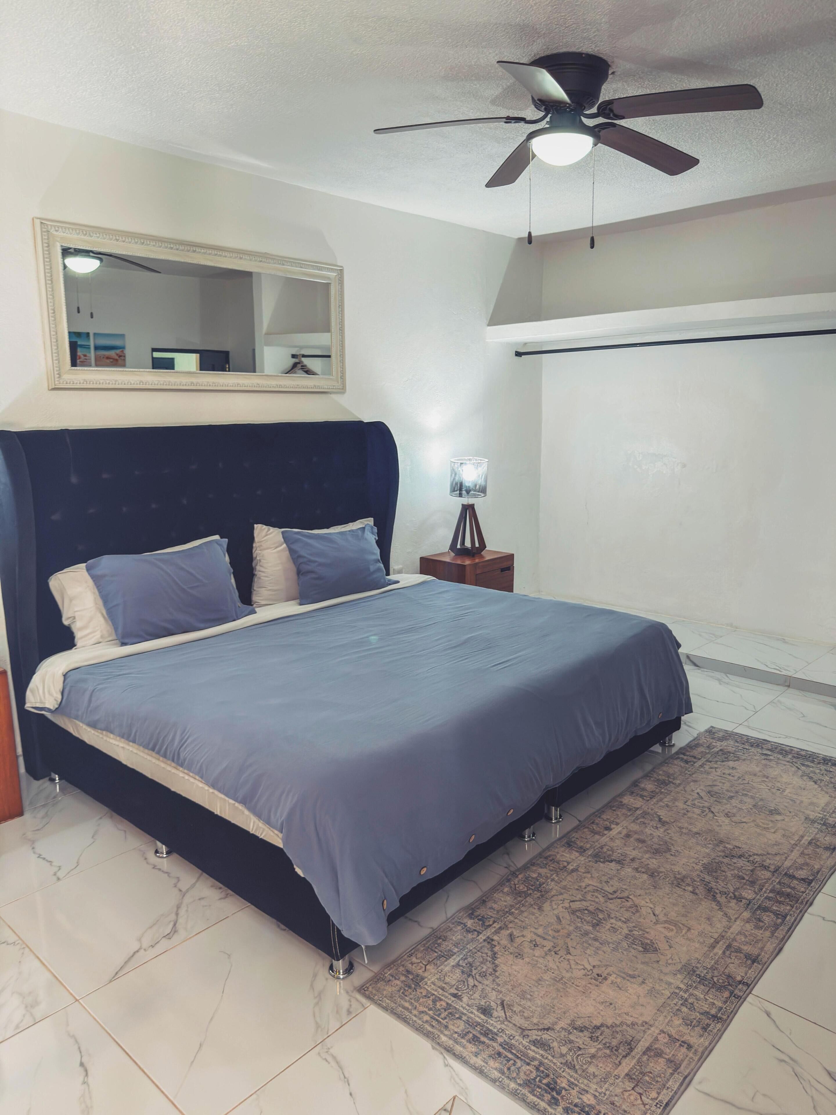 Un dormitorio con cama, ventilador de techo, mesita de noche, lámpara, espejo y alfombra estampada.