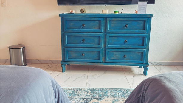 Un dormitorio con una televisión de pantalla plana grande montada en la pared, un aparador azul con varios cajones y una alfombra estampada en el suelo.