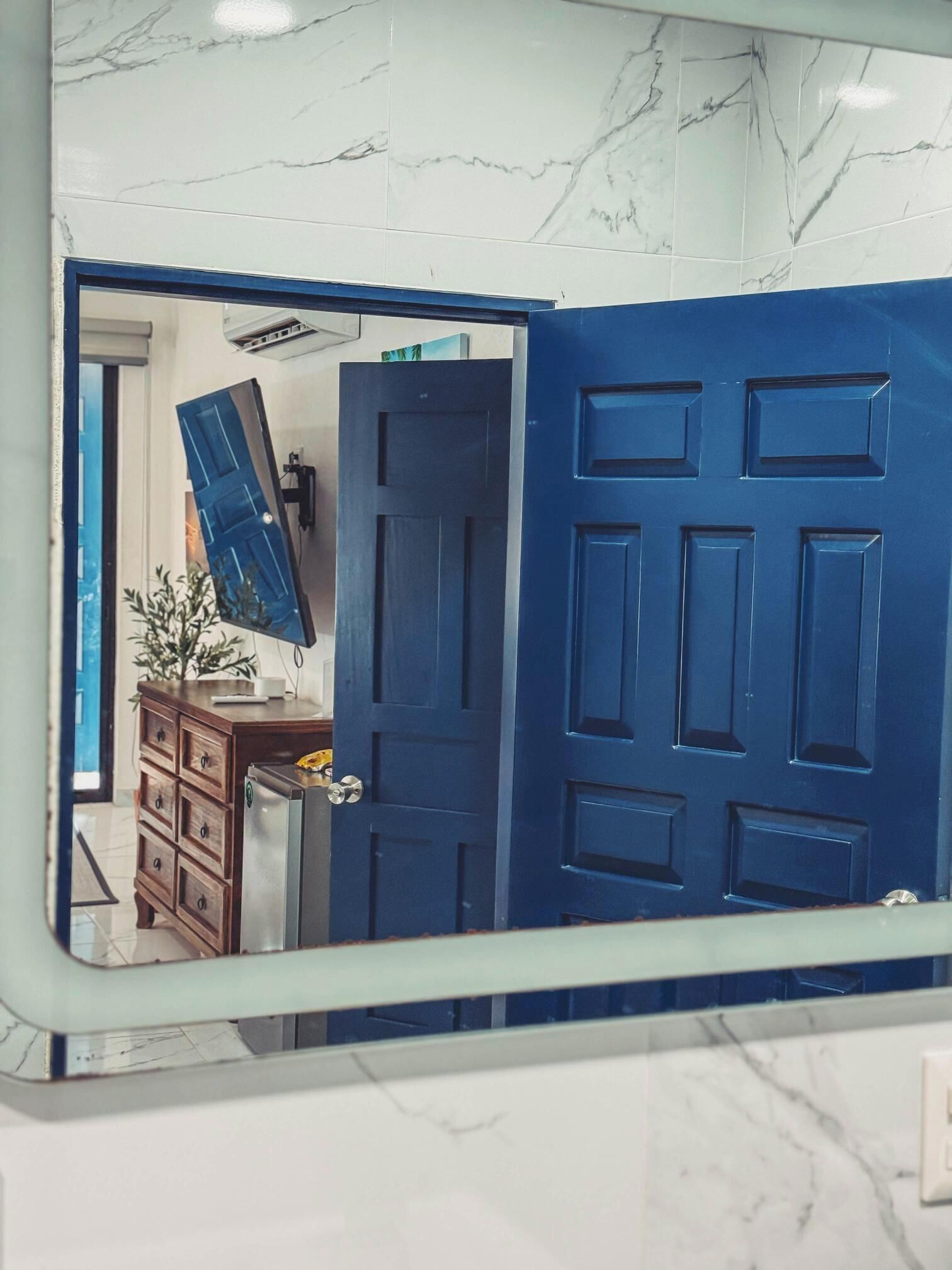 Ein Badezimmer mit Marmorboden, einer blauen Tür und einem Spiegel, der einen Raum mit blauer Tür und einer Holzkommode zeigt.