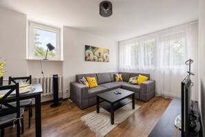 A modern living room with a grey sectional sofa, a coffee table, a dining table with chairs, and a large window with sheer curtains. - Warsaw Praga Apartment — Balcony & Metro Nearby (Warszawa)
