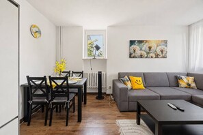 Ein modernes Wohnzimmer mit grauem Sofa, einem Esstisch mit Stühlen, einer Wanduhr und einem Fenster mit Blick auf Bäume.