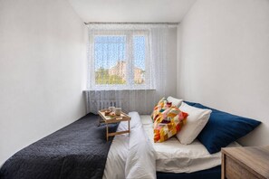 A small, single-bed room with a window, a bedside table, and a tray with items. - Warsaw Praga Apartment — Balcony & Metro Nearby (Warszawa)