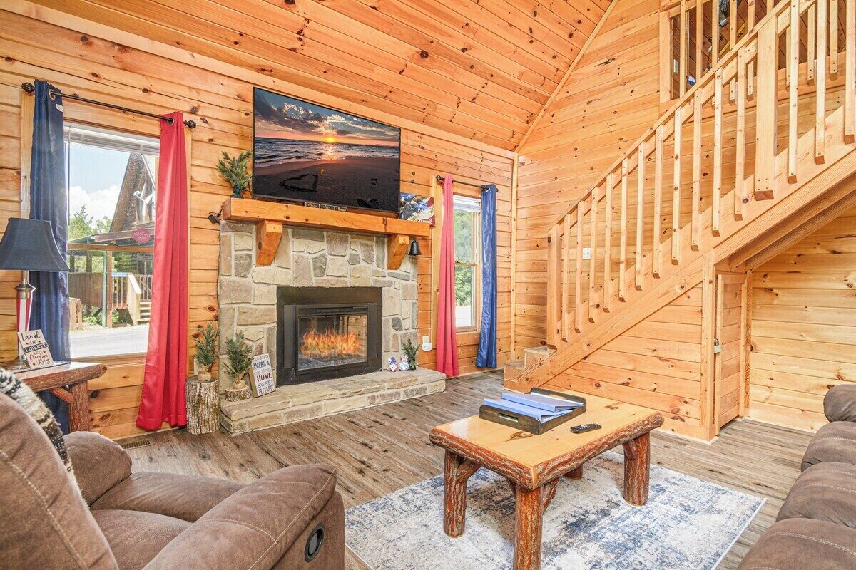 A log cabin interior with a stone fireplace, a television mounted above it, and a staircase leading to an upper level.