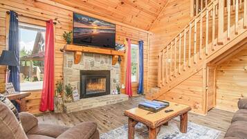 A log cabin interior with a stone fireplace, a television mounted above it, and a staircase leading to an upper level.
