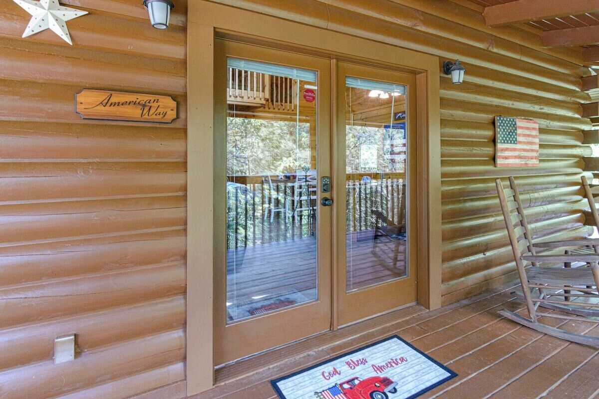 Une cabane en bois avec une enseigne indiquant « American Way », une entrée avec une porte en verre, une terrasse avec des chaises et un drapeau américain.