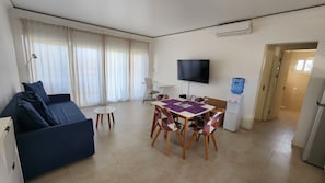 Ein Wohnzimmer mit einer Couch, einem Esstisch mit Stühlen, einem Flachbildfernseher und einem Wasserspender.