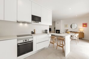 A modern kitchen with white cabinetry, a built-in oven, a microwave, and a dining area with stools. - Casa Floresta - Private Pool Retreat (Óbidos)