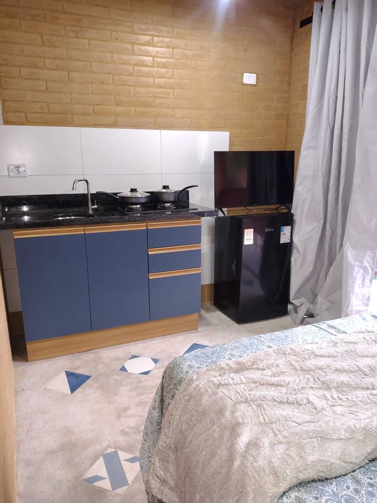 Eine kleine Küche mit Herd, Spüle und Kühlschrank, ein Bett mit gemustertem Überwurf und eine Ziegelwand.