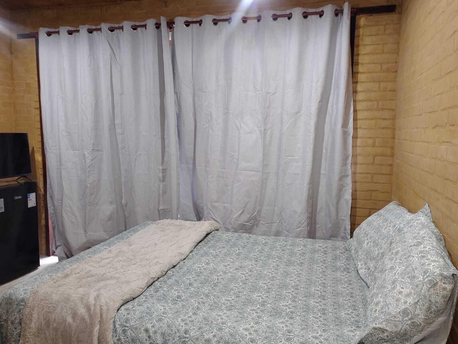 Una cama individual con una colcha estampada, una manta blanca y una almohada en una habitación con pared de ladrillo y cortina blanca.