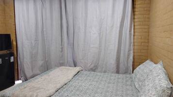 Una cama individual con una colcha estampada, una manta blanca y una almohada en una habitación con pared de ladrillo y cortina blanca.