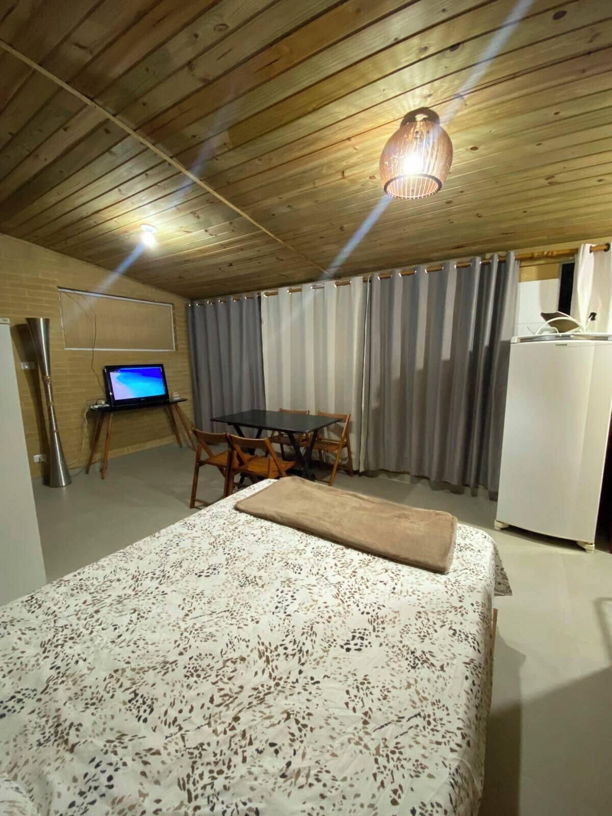 A room with a bed, a desk with a computer, a dining table with chairs, and a refrigerator.
