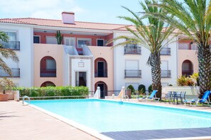 A swimming pool with lounge chairs, palm trees, and a building with balconies and arched doorways. - Blue Horizon, Praia del Rey apartment B9 (Óbidos)