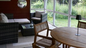 A room with a ceiling fan, a round mirror, a sofa, a dining table with chairs, and large windows offering a view of greenery.