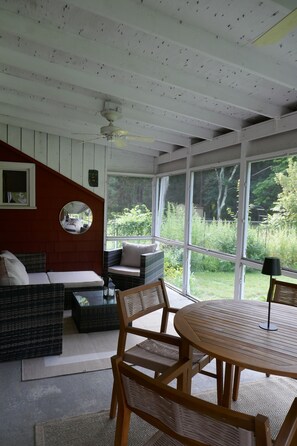A room with a ceiling fan, a round mirror, a sofa, a dining table with chairs, and large windows offering a view of greenery.