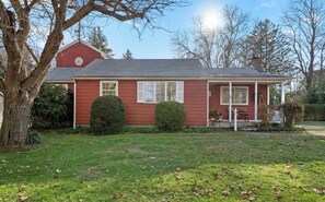 A red single-story house with a front porch, a large tree to the left, and a well-maintained lawn.