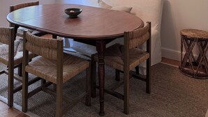 A dining area with a round table, four chairs, a hanging light fixture, a window with blinds, a wall-mounted shelf with decorative items, and a framed floral artwork.