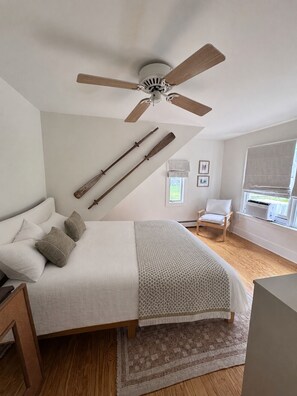 Un dormitorio con cama, ventilador de techo, vigas de madera, ventana con persianas, una silla y un radiador.