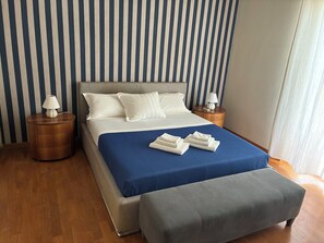 Ein Bett mit blauer Bettdecke, weißen Kissen und einer grauen Sitzbank. Es gibt zwei Nachttische mit Lampen.