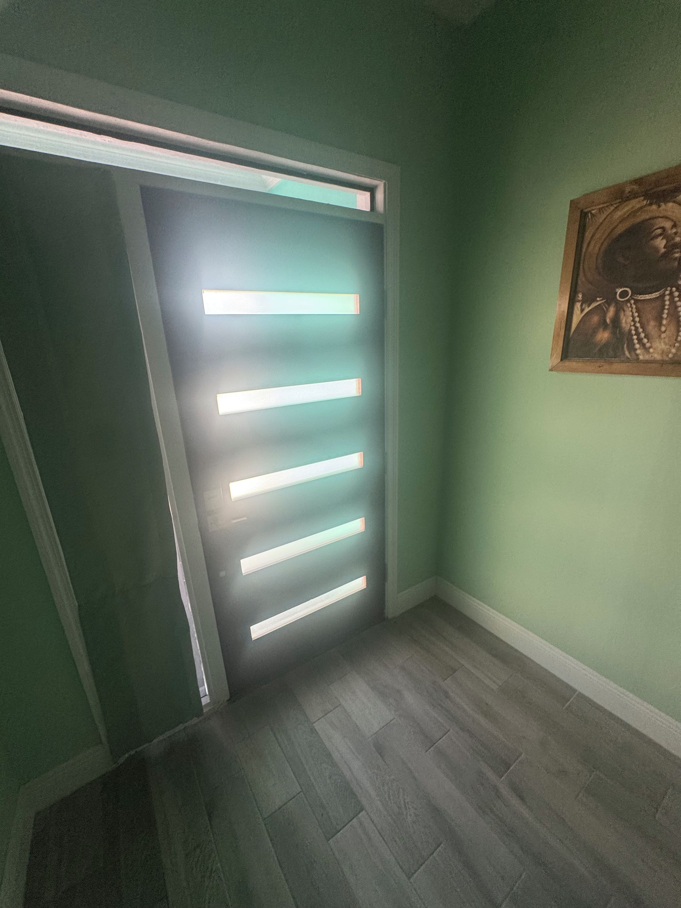 A hallway with a door illuminated by horizontal light strips, a wooden floor, and a framed picture on the wall.