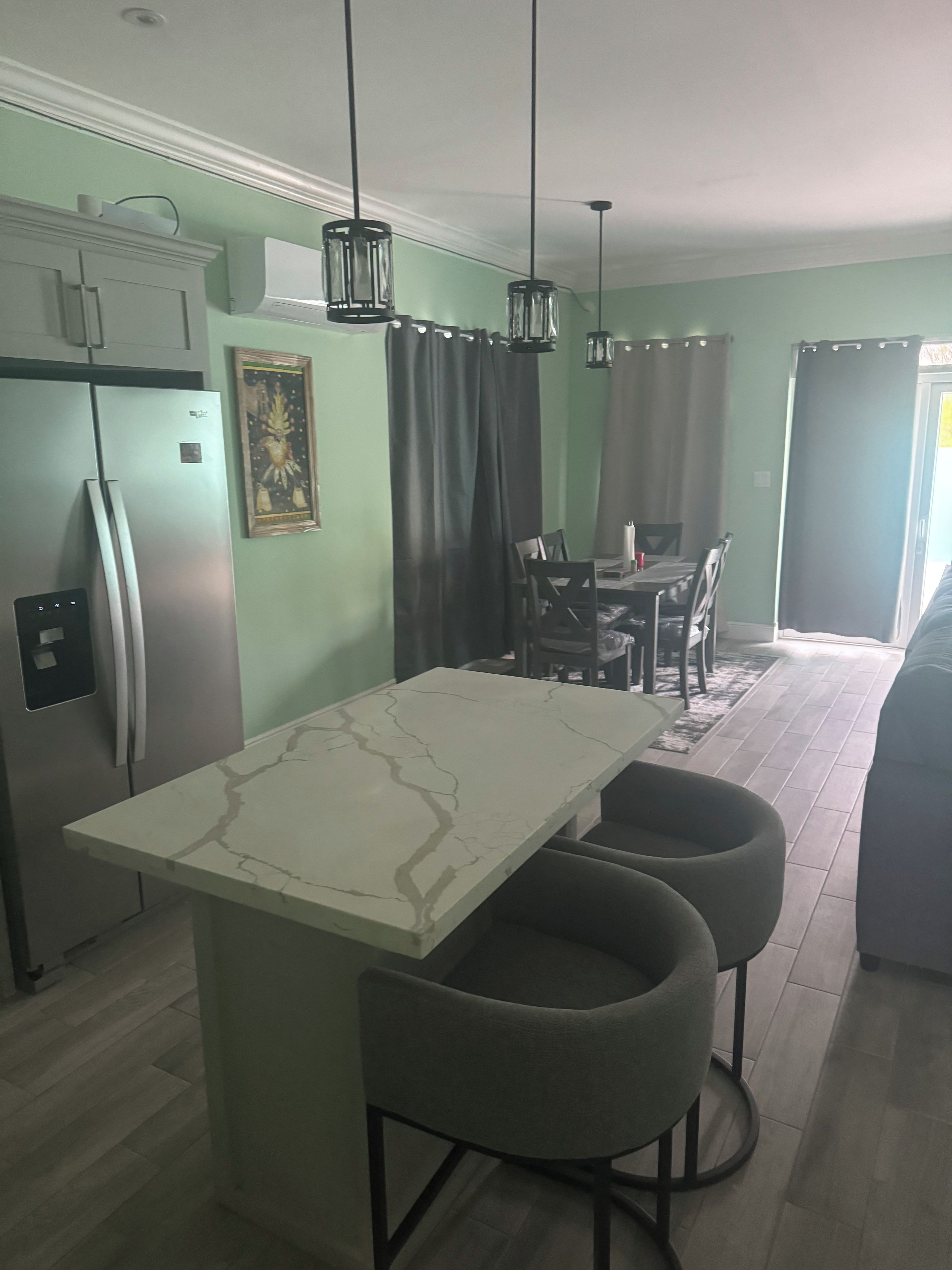 A kitchen with a marble countertop, a refrigerator, pendant lights, and a dining area with chairs and a table.