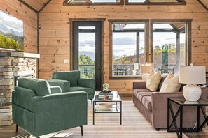A cozy cabin interior with a stone fireplace, green and brown sofas, a coffee table, and large windows offering a view of the outdoors. - Hot Tub + Fire Pit | 3BR Cabin Near Dollywood (Sevierville)