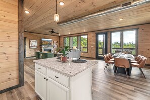 A spacious kitchen with a central island, dining area with chairs, and large windows offering views of the outdoors. - Hot Tub + Fire Pit | 3BR Cabin Near Dollywood (Sevierville)