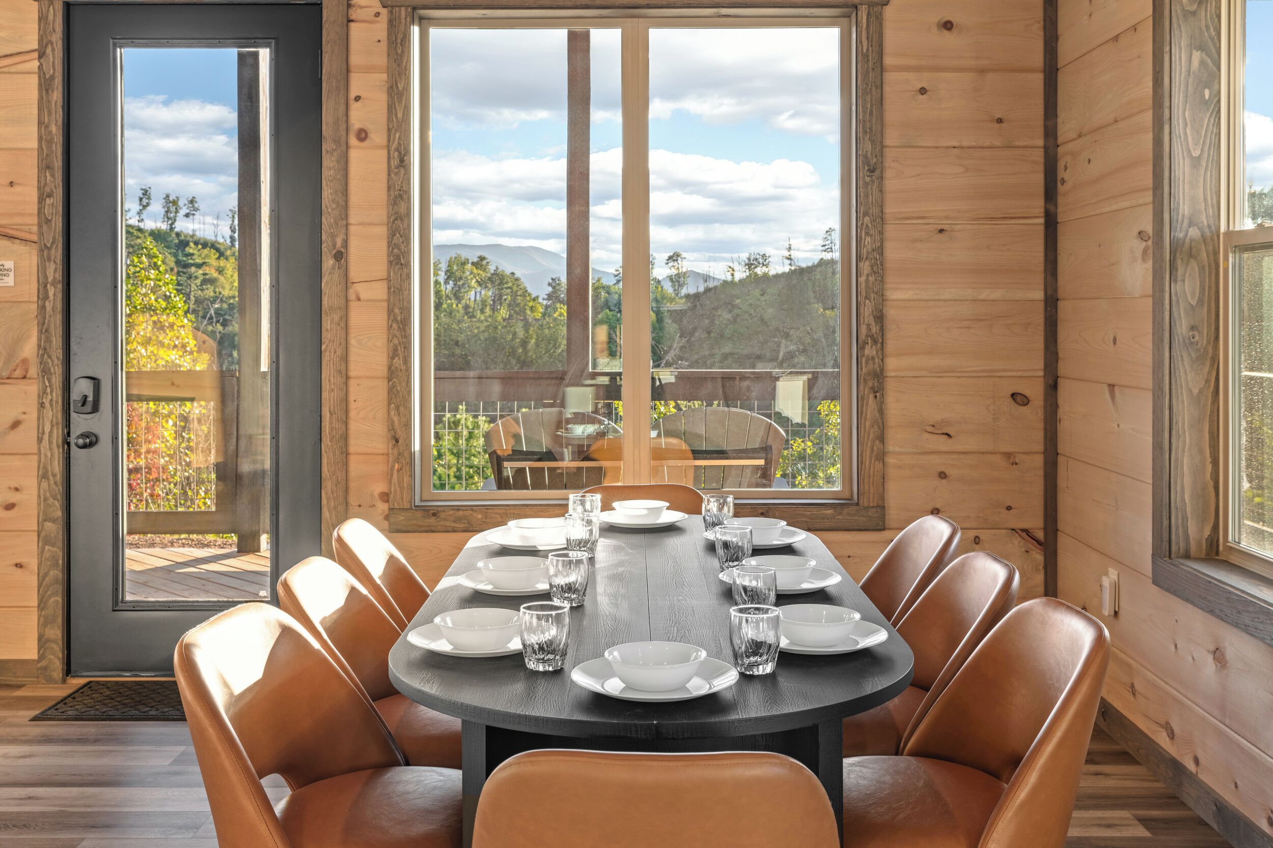 A dining area with a round table set for six, surrounded by leather chairs, with a view of a mountainous landscape through large windows.