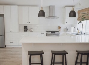 Una cucina moderna con mobili bianchi, un'isola centrale con sgabelli, illuminazione a sospensione e una cappa aspirante in acciaio inossidabile.