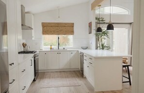 A modern kitchen with white cabinetry, a central island, and a large window with a wooden blind. - Modern 3 Bedroom house in Sacramento w/ Private Sauna (Antelope)