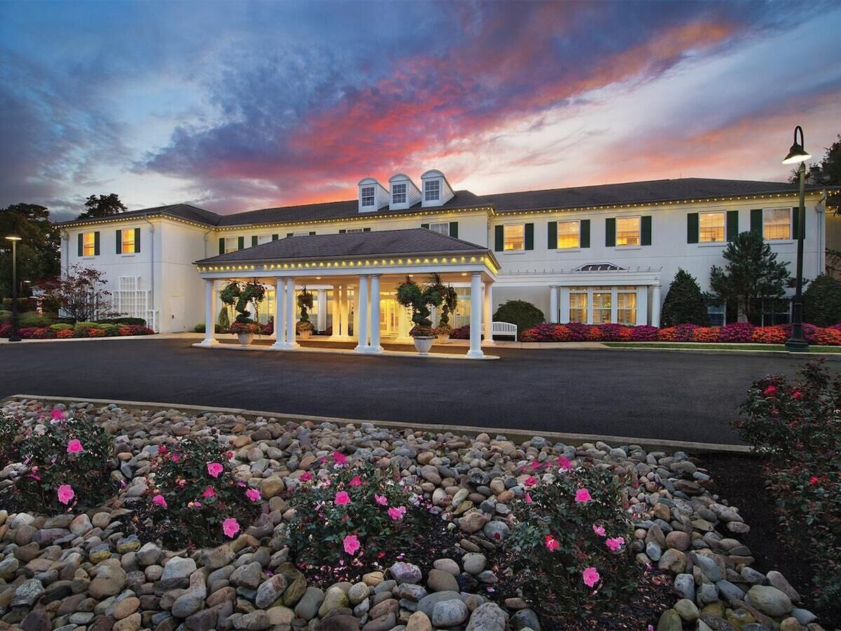 A hotel with a covered entrance, a garden with pink flowers, and a sunset sky.
