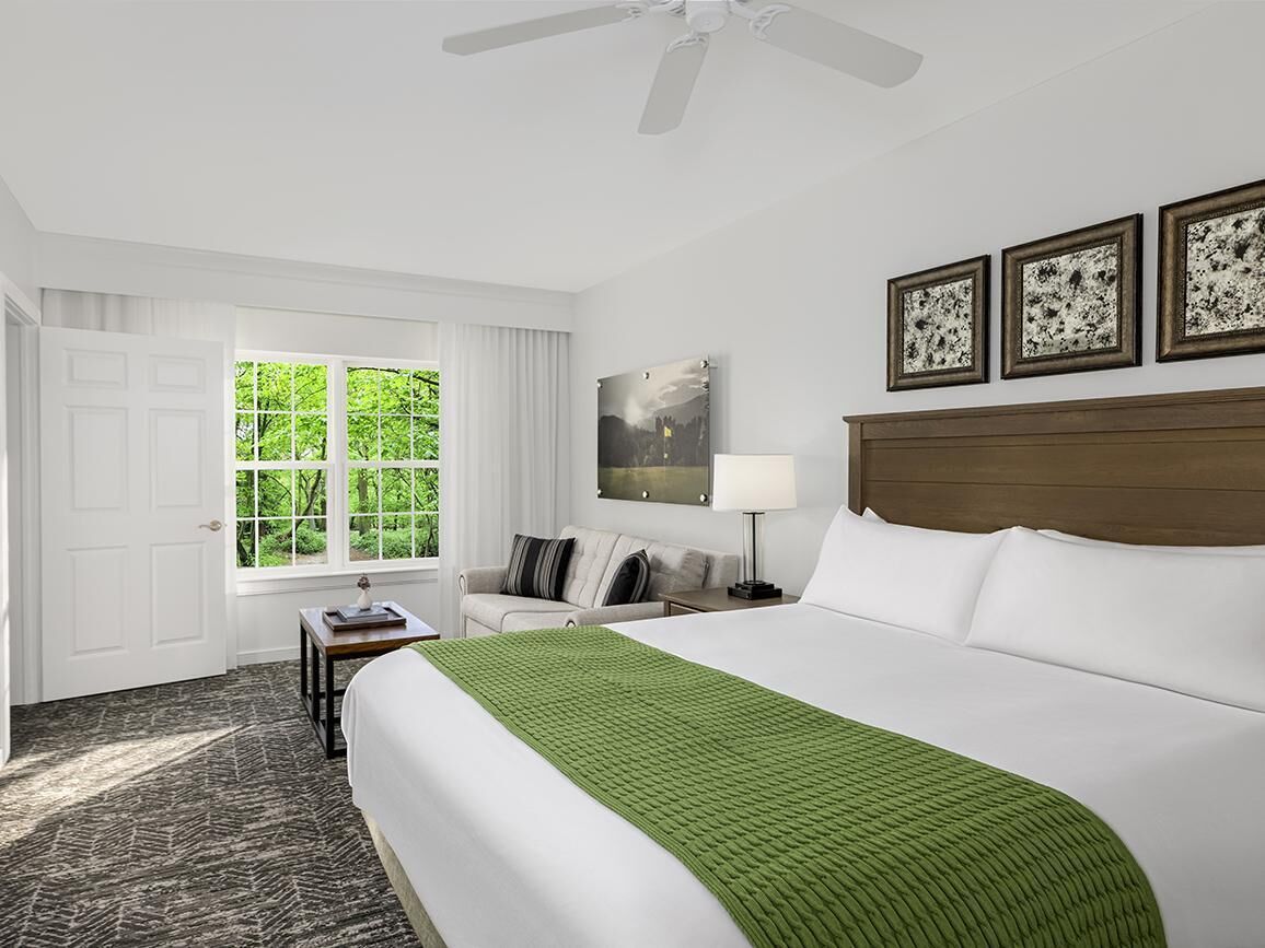 A hotel room with a large bed, a desk, a chair, a window with a view of greenery, and a ceiling fan.