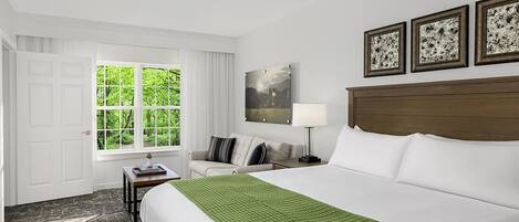 A hotel room with a large bed, a desk, a chair, a window with a view of greenery, and a ceiling fan.