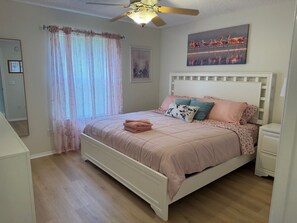 Ein Schlafzimmer mit einem Bett, Deckenventilator, einem Fenster mit Vorhängen, einem Gemälde und einem Nachttisch.