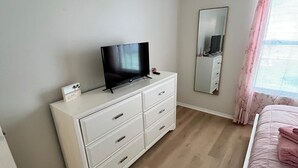 Ein Schlafzimmer mit einem Bett, einem Schminktisch mit Fernseher, einem Spiegel und einem Fenster mit Vorhängen.