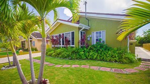 A house with a well-maintained lawn, palm trees, and a variety of plants. - Modern 3BRVilla in Secure Draxhall Gated Community (St Ann)
