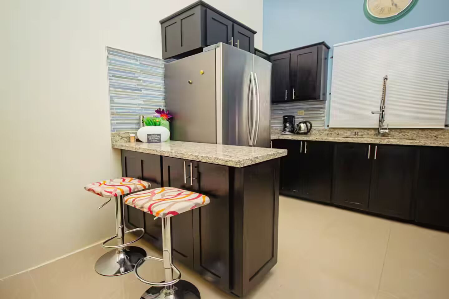 A kitchen with a stainless steel refrigerator, dark cabinets, a granite countertop, and two bar stools with patterned cushions.